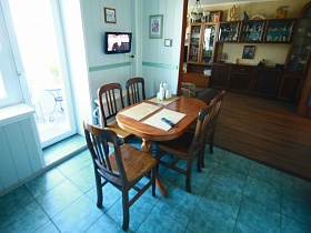 овальный деревянный обеденный коричневый стол с резными ножками на полу с бирюзовой квадратной плиткой у окна с балконной дверью светлой кухни зонированной комнаты квартиры оператора