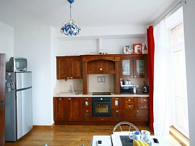коричневая мебельная стенка со встроенной газовой плитой, белый холодильник у белых стен кухни с голубой цветочной люстрой на потолке евро квартиры