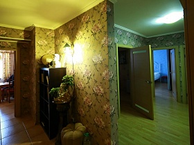 открытый шкаф с полочками и гипсовым бюстом наверху, круглая подставка с фонтаном и бежевый пуфик у стены с ярким светильником в прихожей современной трехкомнатной актерской квартиры