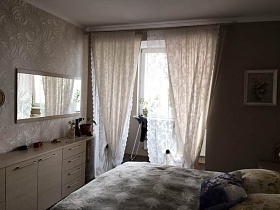 серое покрывало и подушки на большой кровати, прямоугольное зеркало над комодом у бежевых штор на застекленную лоджию современной квартиры