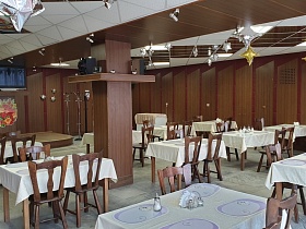 телевизор и колонки на полках вокруг квадратных колон в зале столовой с сервированными столиками на полу с квадратной серой плиткой