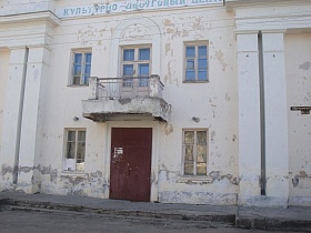 фасад светлого двухэтажного здания старого клуба времен СССР, требующего ремонта с открытым балконом над входной дверью на широком крыльце