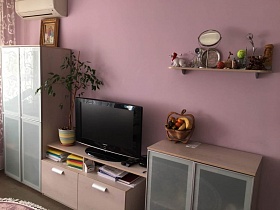 кремовая мебельная горка с иконой на высоком шкафу, комнатным цветком и телевизором на открытой полке и ваза с фруктами на небольшом шкафу у розовой стены просторной гостиной современной двухкомнатной квартиры на 6 этаже