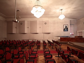 сьемочная камера, большая подвесная люстра на цепях с многочисленными плафонами на потолке светлого актового зала с рядами красных секционных кресел на паркетном полу времен СССР