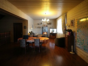камин у стены с полками,телевизор на тумбочке, мягкое кресло и овальный стол в зоне отдыха гостиной большого деревянного дома