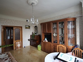 телевизор внутри коричневой мебельной стенки с посудой на полках за стеклянными дверцами в гостиной квартиры сталинки