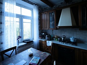 коричневая мебель с голубой мелкой плиткой на рабочей стенке в кухне с прозрачными  гардинами  на окне в семейной трехкомнатной квартире
