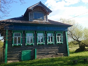деревянная скамейка на пеньках, старое поваленное дерево на открытом участке у деревянного дома на два хозяина с зелеными резными наличниками на окнах, зеленой дверцей подпола, синим карнизом под крышей