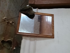 комуфляжная кепка на углу зеркала в деревянной рамке в углу комнаты, требующей ремонта дома заброшенной деревни