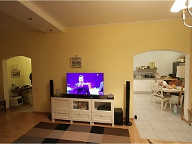 небольшая картина на стене, телевизор на черной поверхности белой тумбы , колонки у стены, между арочными дверными проемами смежных комнат трехкомнатной квартиры