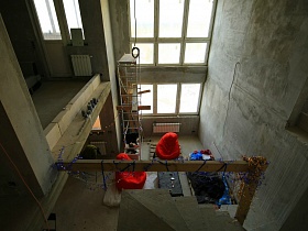 общий вид на просторную двухэтажную квартиру недостроенного пентхауса молодежи с подручным столом, креслами-мешками вокруг, лежаком из деревянных поддонов, строительной лестницей-помостом на бетонном полу первого этажа
