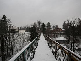 снег на длинном подвесном пешеходном мосту с перилами над домами на крутом берегу реки