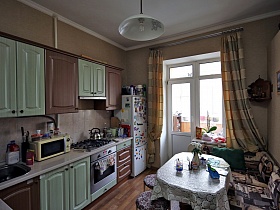 общий вид просторной кухни с угловым диваном вокруг обеденного стола с белой скатертью, мебельной стенкой с белыми и коричневыми дверцами семейной квартиры советского времени