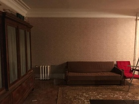 коричневая мебельная стенка со стеклянными дверцами, мягкий складной коричневый диван, красное кресло с подлокотниками на полу с коричневым цветным ковром гостиной с бежевыми стенами квартиры N6