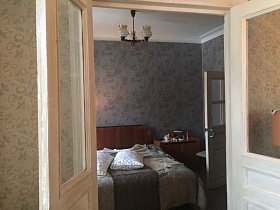 подвесная люстра со стеклянными плафонами на белом потолке спальни с серыми обоями на стенах, большая деревянная кровать с цветным покрывалом из открытой двери кв.24