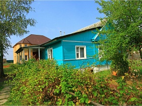 голубая деревянная старая дача СССР с открытой верандой под навесом и желтое кирпичное двухэтажное здание с коричневой крышей на зеленом участке за деревянным забором у поля
