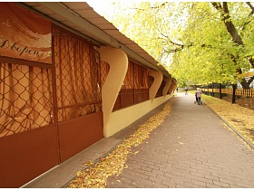опавшие желтые листья на длинной алее вдоль деревьев у высокого металлического забора и летнего кафе на озере