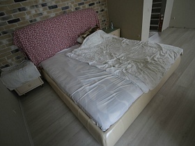 большая кровать с белым постельным, красным одеялом на спике, прикроватными тумбочками у стены , выложенной разноцветным кирпичом