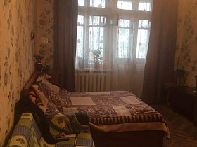 диван с квадратным зеленым покрывалом, большая кровать с коричневым покрывалом, тумбочка, ковер на полу спальной комнаты с бежевыми цветными обоями и коричневыми шторами на окне квартиры СССР