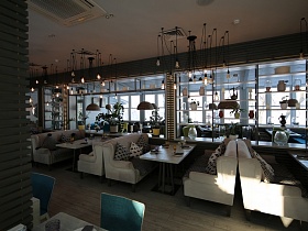 потолочные люстры в стиле хай-тек над белыми столиками с мягкими белыми диванами с подушками у длинного прозрачного стеллажа с полками в светлом зале ресторана