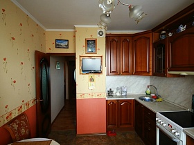 коричневая кухня со светлой столешницей и рабочей поверхностью, картины на бежевой стене с рыжими панелями в яркой современной трехкомнатной квартире с комнатой бабушки