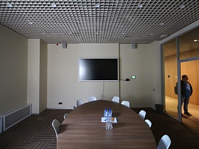 белый плоский телевизор на светлой стене переговорной комнаты с большим овальным столом по центру и стеклянной стеной