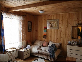 мягкий диван с леопардовым покрывалом и подушками и белая тубмочка у стены с картинами и пано в гостиной дероевянной дачи СССР