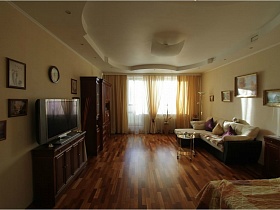 телевизор на комоде,коричневая мебельная стенка, двухцветный диван с подушками и картины на стенах гостиной простой средней двухкомнатной квартиры с балконами