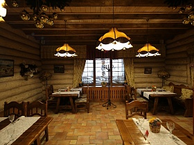 картины и деревянные резные полки с посудой на бревенчатых стенах, торшеры с желтыми абажурами в углах комнаты, напольная вешалка у большого окна уютной комнаты просторного ресторана в купеческом стиле