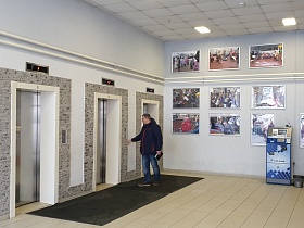два терминала у светлой стены с большими фотографиями под стеклом в просторном холле с лифтами