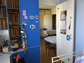 наклейки, заметки на голубой дверце углового компьютерного стола с книгами на полках, перекидной календарь на белой межкомнатной двери в голубую спальную комнату трехкомнатной квартиры