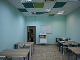 белые пластиковые стулья со спинкой у деревянных столов светлой учебной комнаты с голубыми стенами, синими и зелеными квадратными вставками на белом потолке