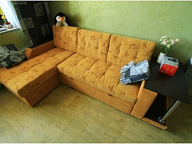 пингвинчик и квадратные подушки на желтом угловом диване с боковым столиком в гостиной двухкомнатной квартиры новостроя