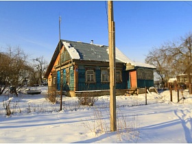 неухоженный деревянный домик, окрашенный в голубой цвет за забором из сетки, на участке с нетронутым белым снегом в деревне