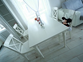 белые стулья со спинкой вокруг прямоугольного белого стола с фруктами и подсвечниками на поверхности напротив окна в зоне кухни двухкомнатной квартиры