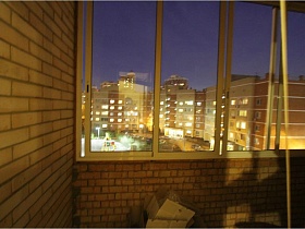 современные высотные соседние дома из окна застекленной лоджии с кирпичными стенами и картонными коробками на полу простой трехкомнатной квартиры