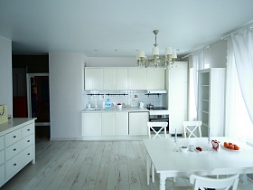 белая вытяжка над встроенной газовой плитой в белой кухне с кухонными принадлежностями на белой столешнице зонированной кухни светлой современной квартиры в новостройках