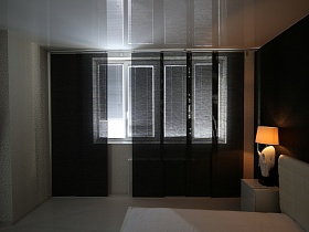 белые горизонтальные жалюзи на стеклах и черные векртикальные на большом окне черно белой спальной комнаты современной квартиры