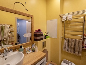 зеркало в деревянной рамке над деревянной столешницей с белой раковиной,цветной короб и косметические принадлежности на поверхности встроенного шкафа, сушилка для белья над полотенцесушителем у желтых стен ванной комнаты сталинской квартиры