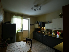 обеденный стол со скатертью, серый холодильник у стены с маками, черно-белая мебельная стенка в светлой кухне с салатовой гардиной на окне с балконной дверью
