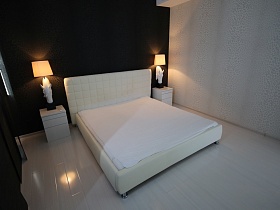 белые настольные лампы с изображением животных с абажурами на прикроватных тумбочках у большой кровати на металлических ножках с белым покрывалом в черно белой спальне