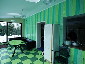 мебель на полу закрытой яркой летней кухни таунхауса с шахматной двухцветной плиткой, салатовой плиткой на стенах и подвесными люстрами на белом потолке