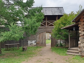 деревянный бревенчатый дом без покраски с открытым крыльцом с перилами под навесом на большом дворе за высоким забором и арочными воротами в русской деревне
