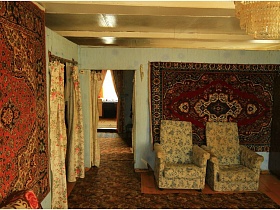 ковры на стенах, цветные шторы на дверях, старая люстра, большие мягкие кресла у стены гостиной с ковровыми дорожками на полу жилого дома на селе