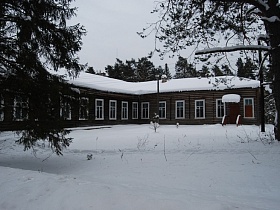 большой заснеженный двор с одноэтажной старинной деревянной угловой школой темно-коричневого цвета с шапкой снега на крыше