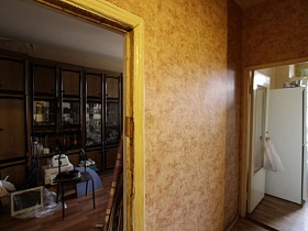 коричневая мебельная стенка советских времен в зале и белый холодильник на кухне через открытые двери в прихожей бедной квартиры в жилом доме