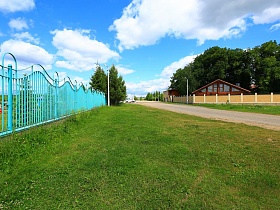 асфальтированная дорога между желтым бетонным забором с колоннами и голубым металлическим фигурным забором в живописном месте с сосновым лесом и церквью
