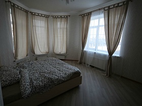 большая бежевая кровать с цветным постельным, бежевые шторы на окнах светлой спальной комнаты кирпичного двухэтажного дома под съем