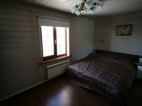 картина над коричневой большой кроватью с полосатым покрывалом у окна с жалюзи в спальне просторного деревянного дома