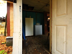 открытая белая дверь веранды с белым холодильником и машинкой на неокрашенном деревянном полу бедного жилого домика у пруда